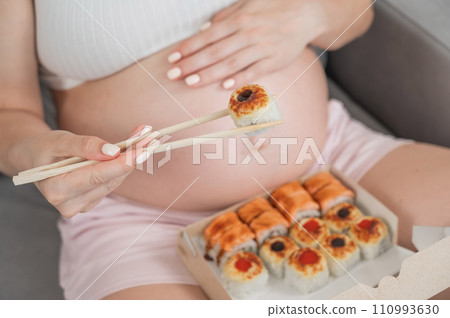 A pregnant woman sits on the sofa and eats rolls from a box. Food delivery. Close up of belly. A pregnant woman sits on the sofa and eats rolls from a box. Food delivery. Close up of belly. 110993630