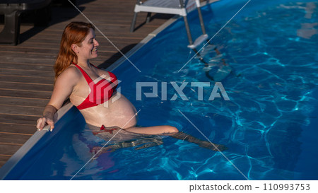 A red-haired pregnant woman in a red bikini is resting in the pool, leaning on the side. A red-haired pregnant woman in a red bikini is resting in the pool, leaning on the side. 110993753