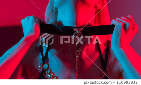 Blonde woman in leather mask putting on collar in bedroom with red blue neon light. Blonde woman in leather mask putting on collar in bedroom with red blue neon light. 110993932