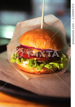 Burger with cheese on wax paper on plastic tray on wooden table. Fresh tasty burger on dark background. Close up. High quality photo 110994355