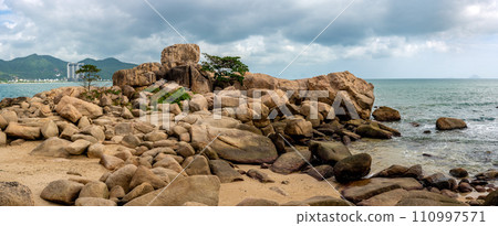 Coastal waves crash against monumental granite boulders lining shore. majestic seascape features vast expanse of rocks, under turbulent, cloud-laden sky, untamed natural panorama 110997571