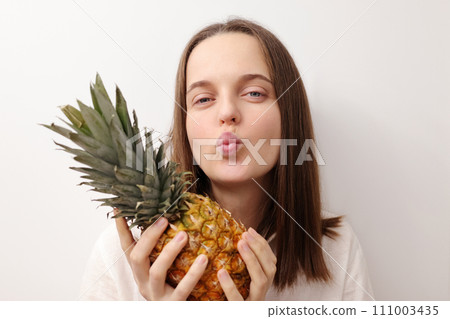 Romantic beautiful joyful Caucasian woman holding pineapple isolated over gray background promoting healthy eating with fresh vegetables sending air kissing Romantic beautiful joyful Caucasian woman holding pineapple isolated over gray background promoting healthy eating with fresh vegetables sending air kissing 111003435