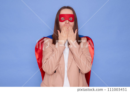 Shocked brown haired woman wearing superhero costume posing isolated over blue background making mistake covering mouth with hands sees something astonishing Shocked brown haired woman wearing superhero costume posing isolated over blue background making mistake covering mouth with hands sees something astonishing 111003451