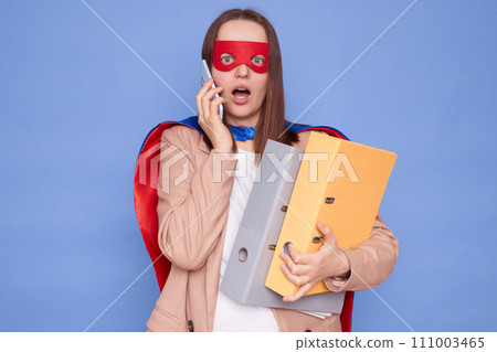 Shocked brown haired woman wearing superhero costume holding documents in folders posing isolated over blue background talking on mobile phone hearing information about work deadline Shocked brown haired woman wearing superhero costume holding documents in folders posing isolated over blue background talking on mobile phone hearing information about work deadline 111003465