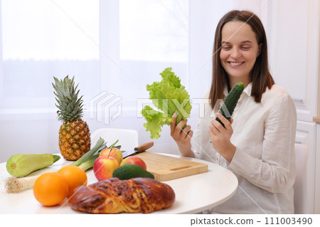 Cheerful positive attractive Caucasian brown haired woman sitting at kitchen table with fruit and vegetables holding cucumber and green lettuce leaves in hands 111003490