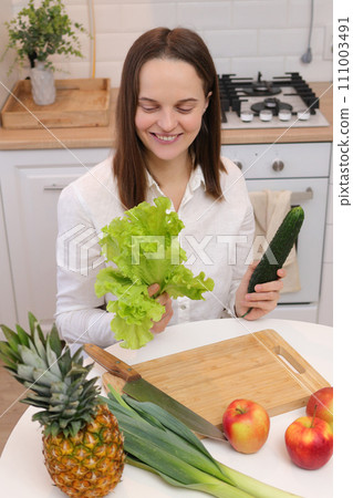 Delighted satisfied joyful Caucasian brown haired woman sitting at kitchen table with fruit and vegetables holding cucumber and green lettuce leaves in hands organic ingredients for salad 111003491