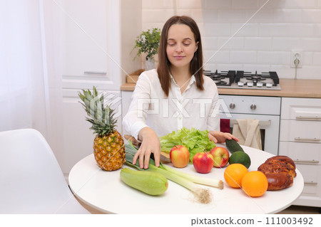 Healthy lifestyle. Raw vegan. Domestic kitchen. Nutritious dinner. Caucasian brown haired woman sitting at kitchen table with fruit and vegetables cooking organic meals at home 111003492
