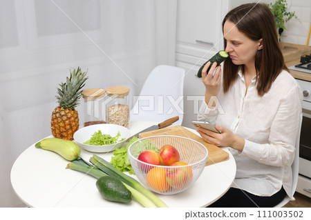 Confused puzzled Caucasian brown haired woman sitting at kitchen table with fruit and vegetables smelling not fresh cucumber from refrigerator or freezer 111003502