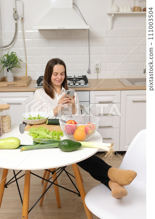 Satisfied smiling delighted Caucasian brown haired woman sitting at kitchen table with fruit and vegetables relaxing with smartphone in hands searching veggies receipts online Satisfied smiling delighted Caucasian brown haired woman sitting at kitchen table with fruit and vegetables relaxing with smartphone in hands searching veggies receipts online 111003504