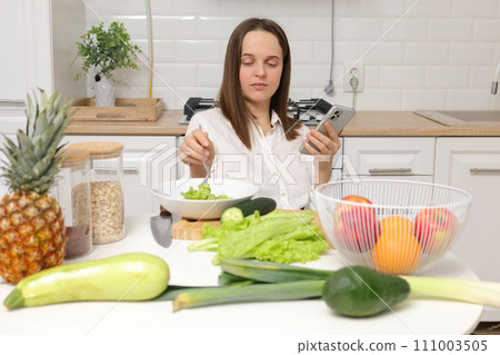 Confident Caucasian brown haired woman sitting at kitchen table with fruit and vegetables using smartphone while eating fresh green salad with lettuce leaves 111003505