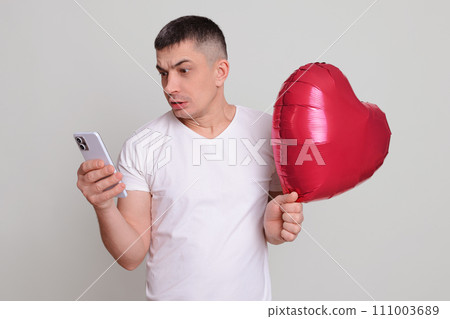 Confused shocked Caucasian brunette man holding red helium balloon in heart shape isolated over gray background using mobile phone reading online message from girlfriend Confused shocked Caucasian brunette man holding red helium balloon in heart shape isolated over gray background using mobile phone reading online message from girlfriend 111003689