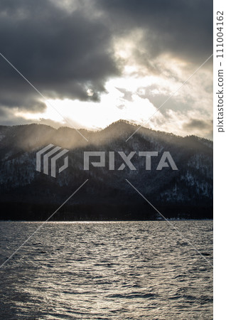 Dramatic scenery of a mountain lake where the light of the setting sun is blocked by clouds and hits the mountains. Lake Kussharo in Hokkaido. 111004162
