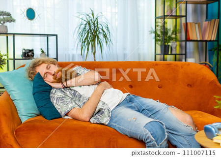 Tired Caucasian redhead man lying down in bed taking a rest at home, napping, falling asleep on sofa 111007115