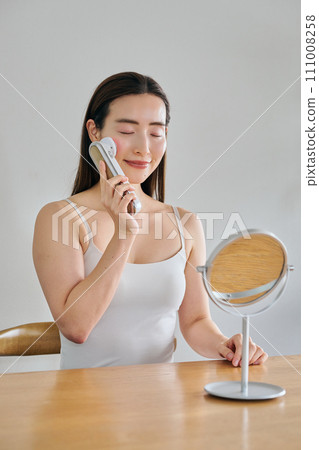 A woman in her 30s with long hair doing skin care using a facial beauty device 111008258