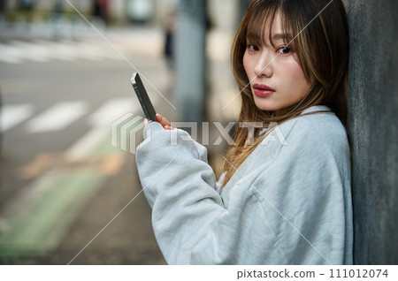 Young woman leaning against the wall and operating a smartphone Young woman leaning against the wall and operating a smartphone 111012074