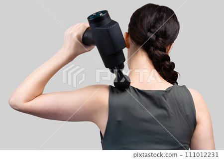 Back view of fit woman gives herself massage on upper arm with percussion massager-gun with ball nozzle. Gray studio background. Therapy for joint pain relief Back view of fit woman gives herself massage on upper arm with percussion massager-gun with ball nozzle. Gray studio background. Therapy for joint pain relief 111012231