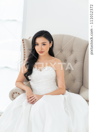 A beautiful, smiling Southeast Asian bride in white wedding gown sitting on sofa. Wedding beauty portrait in asia concept 111012372