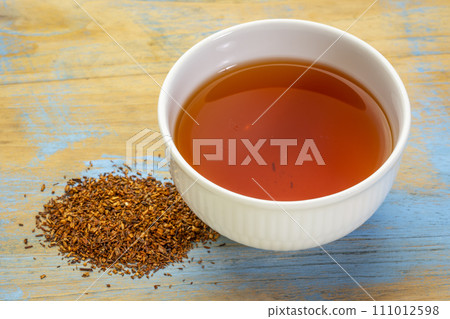 rooibos red tea  -  a white cup of a hot drink and loose leaves on grunge wood background, tea made from the South African red bush, naturally caffeine free 111012598