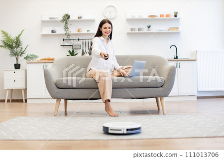 Lady switching on robot vacuum cleaner via remote control sitting on sofa. Lady switching on robot vacuum cleaner via remote control sitting on sofa. 111013706