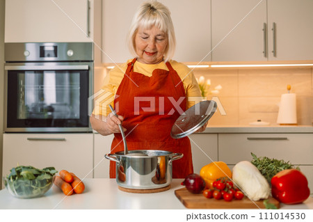 Senior 60s woman standing looking and smelling tasting fresh delicious from soup in a pot with steam at white interior kitchen Senior 60s woman standing looking and smelling tasting fresh delicious from soup in a pot with steam at white interior kitchen 111014530