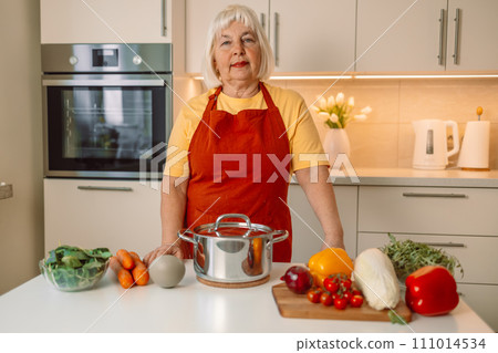 Senior 60s woman standing looking and smelling tasting fresh delicious from soup in a pot with steam at white interior kitchen Senior 60s woman standing looking and smelling tasting fresh delicious from soup in a pot with steam at white interior kitchen 111014534