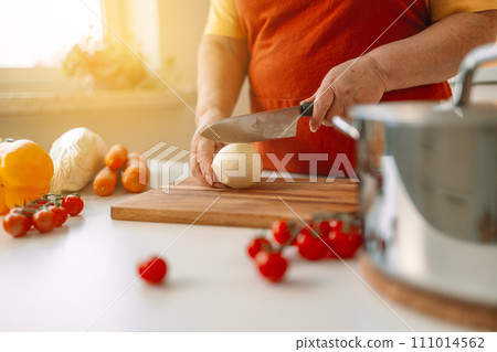 Senior housewife slices onion into pieces on a wooden chopping board in a modern kitchen. Female cutting fresh raw onion on wooden background 111014562