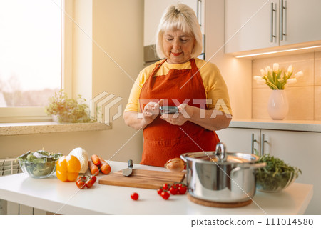 Woman housewife holds phone using cooking apps websites search recipes, chatting with friend during food preparation. Female cooking vegetables in cozy wooden kitchen n 111014580