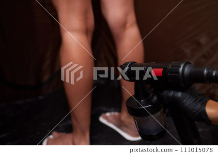 Cosmetologist using airbrush for spray tan apply to young woman body in beauty salon. 111015078