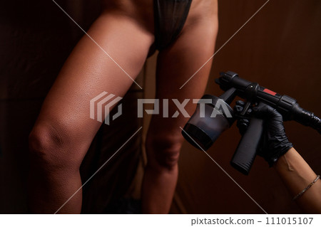 Cosmetologist using airbrush for spray tan apply to young woman body in beauty salon. 111015107