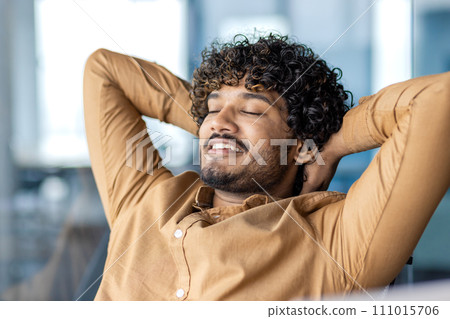 Portrait of relaxed latino guy in camel shirt leaning on chair back and holding hands behind head with wide smile. Productive entrepreneur having break from working on startup with impressive profit. Portrait of relaxed latino guy in camel shirt leaning on chair back and holding hands behind head with wide smile. Productive entrepreneur having break from working on startup with impressive profit. 111015706