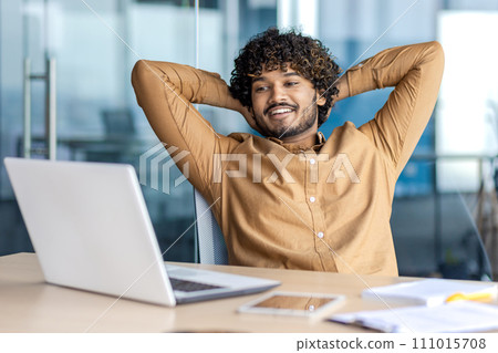 Young indian man in formal clothes stretching back while sitting by desktop and looking at laptop screen with smile. Confident employee feeling no stress and watching result of successful working day. Young indian man in formal clothes stretching back while sitting by desktop and looking at laptop screen with smile. Confident employee feeling no stress and watching result of successful working day. 111015708