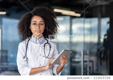 Qualified black woman doctor standing in medical center and holding digital tablet in hands while looking at camera. Serious female physician in uniform ready for meeting patients and giving help. 111015720