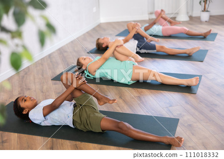 Children doing yoga with group in studio, lying on mat with knee pressed to chest with hands, performing hamstring and low back stretching Ardha Pawanmuktasana Children doing yoga with group in studio, lying on mat with knee pressed to chest with hands, performing hamstring and low back stretching Ardha Pawanmuktasana 111018332