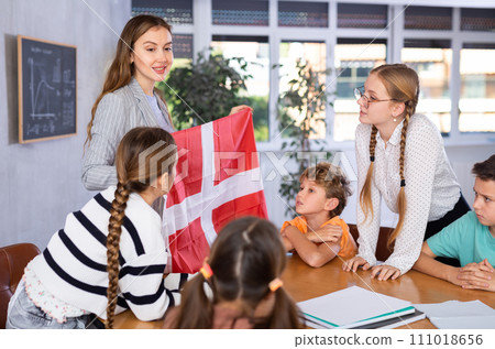 Positive young female teacher explaining culture of Denmark country while showing state flag to schoolchildren preteens in classroom 111018656