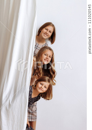 Three girls peek out from behind a curtain against a white wall 111020545