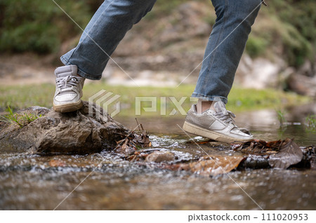 A close-up image of an adventure woman crossing a small nature canal while hiking on the mountain. 111020953
