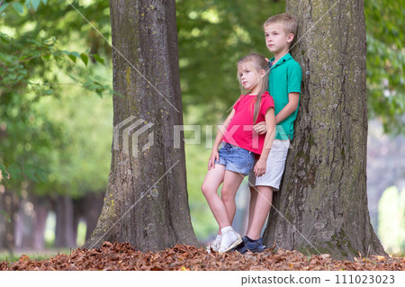 Portrait of two pretty cute children boy and girl standing near big tree trunk in summer park outdoors. 111023023
