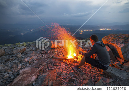 Young tourist man sitting on summer night at bright fire on rocky mountain top under cloudy sky. Young tourist man sitting on summer night at bright fire on rocky mountain top under cloudy sky. 111023058