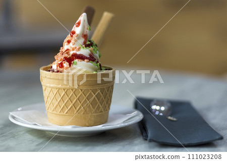 White plate with ice cream dessert in wafer cup with chocolate cookies and creative decoration topping on blurred colorful interior background. 111023208