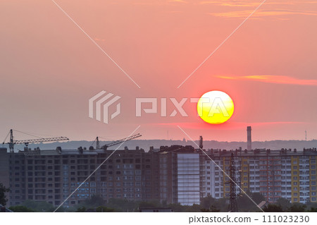 Beautiful sunrise in city suburbs. Big bright white and yellow sun over high apartment building, working tower cranes and houses roofs among green trees. Construction and real property concept. 111023230