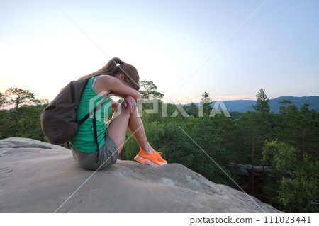 Lonely young woman sitting depressed on hillside trail on warm summer evening. Lost female hiker crying on rocky cliff on wilderness path. Depression and loneliness concept Lonely young woman sitting depressed on hillside trail on warm summer evening. Lost female hiker crying on rocky cliff on wilderness path. Depression and loneliness concept 111023441