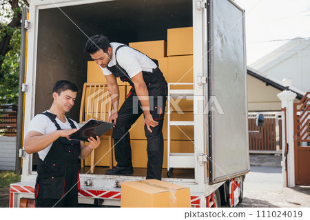 Uniformed workers unload cardboard boxes checking checklist with a clipboard at the truck. Reliable movers guarantee efficient moving and professional service. Moving day concept Uniformed workers unload cardboard boxes checking checklist with a clipboard at the truck. Reliable movers guarantee efficient moving and professional service. Moving day concept 111024019