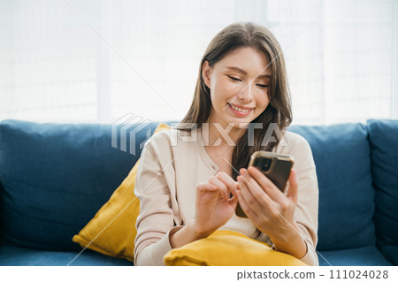 Amid home comfort a woman on a sofa types a message on her phone embracing successful communication. With a smiling face she enjoys technology and relaxation. The woman is relaxing. 111024028