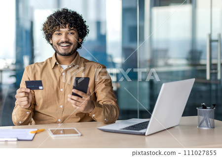Happy indian man with trendy hairstyle sitting by equipped desktop with mobile phone and plastic debit card in hands. Male customer feeling satisfied with getting cashback from online shopping. 111027855