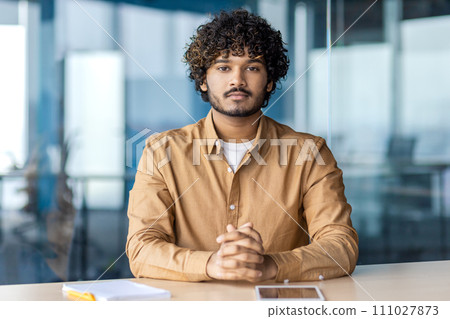 Strict indian businessman sitting with hands folded on table and looking confidently at camera on blurred background. Focused male thinking over company future and pondering over project development. 111027873