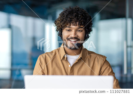 Smiling arab man with curly hair in wireless headset looking at laptop screen while sitting in office. Happy trainee programmer enjoying watching educational webinar for increasing work productivity. Smiling arab man with curly hair in wireless headset looking at laptop screen while sitting in office. Happy trainee programmer enjoying watching educational webinar for increasing work productivity. 111027881