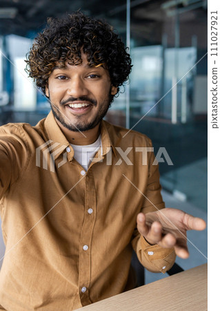 First person view of muslim guy in smart casual clothes having phone video call and smiling while sitting at desktop. Inspired manager showing family new workspace using wireless internet connection. 111027921