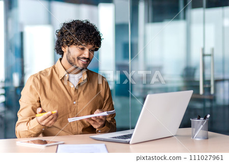 Attentive arab man holding notepad and pen while sitting by table with laptop in modern workplace. Positive freelancer writing down clients wishes and ideas about project realisation via video call. Attentive arab man holding notepad and pen while sitting by table with laptop in modern workplace. Positive freelancer writing down clients wishes and ideas about project realisation via video call. 111027961