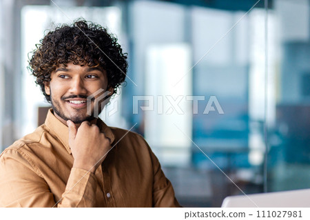 Portrait of muslim male in mustard shirt looking aside and resting head on hand with clenched fist. Smiling man daydreaming and imagining upcoming vacations during working day at modern office. 111027981