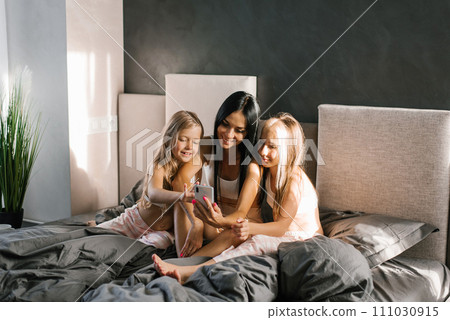 Mom and her two daughters take selfies on a smartphone, relaxing on the bed or video chat. A mother and her children have fun together. 111030915